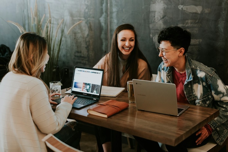 Three colleagues laughing together over laptops at a cafe table